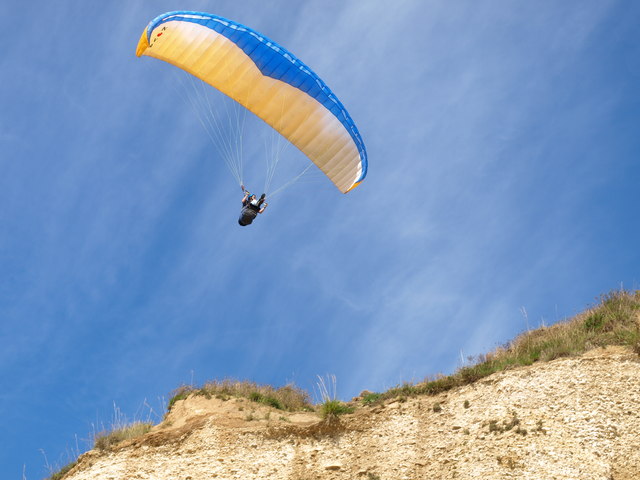 A paraglider using a blue and yellow chute soars over a mountainous desert